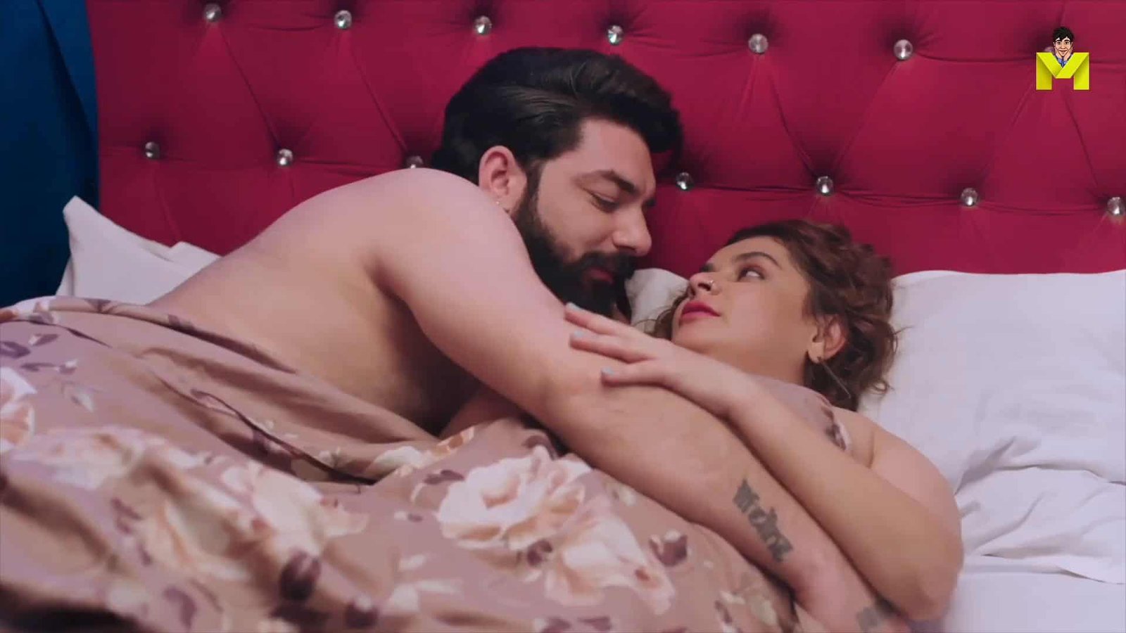 Intimate couple lying in bed, facing each other in a romantic scene with red headboard.